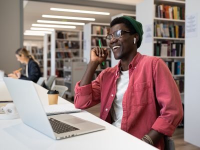 Happy excited African American man student sits in library at table with laptop and laughing